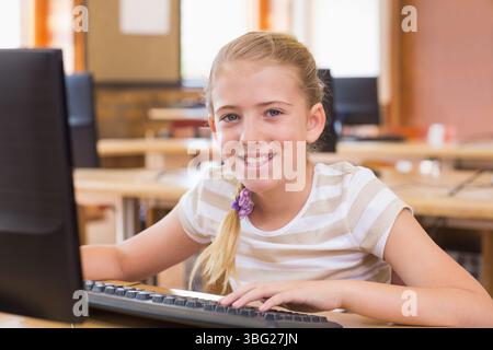Female child student typing on keyboard and smiling at monitor in classroom with bulletin board Stock Photo