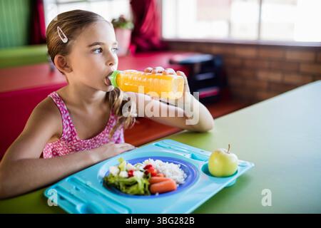 Girl sipping orange juice from bottle in cafeteria, holding blue tray with rice veggies and apple Stock Photo
