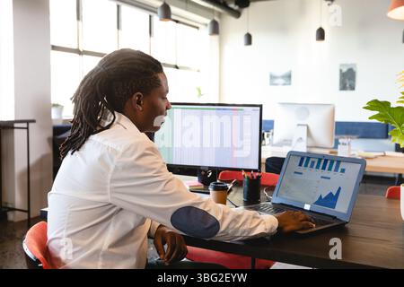 Laptop displaying charts with monitor showing data on open-plan desk with coffee cup and plant Stock Photo