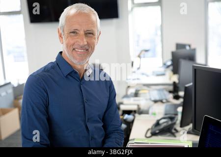 Senior man wearing blue shirt leaning against desk smiling in office with laptop, copy space Stock Photo