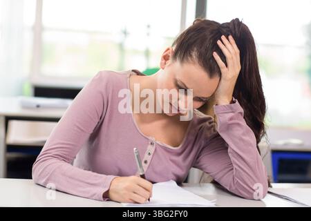 Woman wearing mauve long-sleeve top writing with pen on paper at classroom desk while resting head Stock Photo