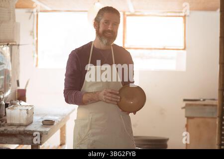 Middle-aged potter wearing apron holding freshly thrown clay bowl near workbench in pottery studio Stock Photo