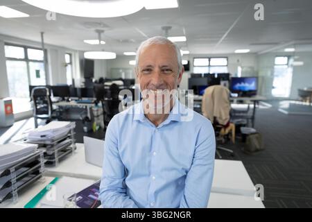 Senior man sitting on white desk in open-plan office smiling with laptop documents glass of water Stock Photo