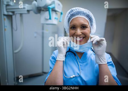 Female nurse pulling down surgical mask with gloved hands in radiology room with X-ray machine Stock Photo