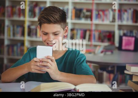 School-age boy sitting at library table holding smartphone reading open book with stacked books Stock Photo