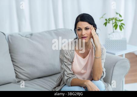 Woman pressing temple on sofa in living room near side table with tissue box, copy space Stock Photo