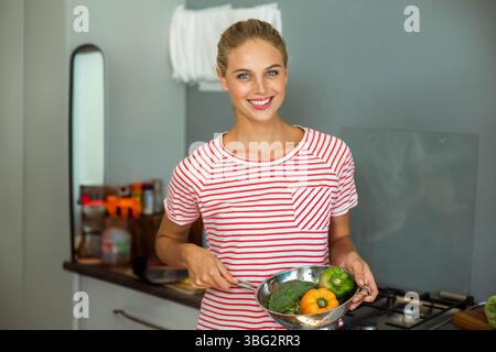 Woman holding metal colander filled with broccoli and bell peppers in kitchen near spice jars Stock Photo
