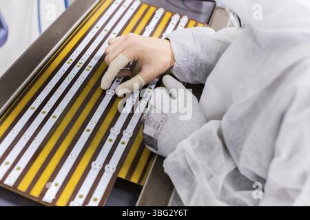 Close-up of a factory worker in a white lab suit and latex gloves, sitting on the chair and installing LEDs in a tv set Stock Photo