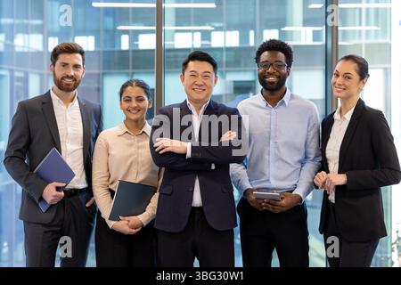 A diverse group of business professionals pose together in a modern office setting, smiling at the camera. Stock Photo