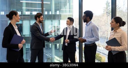 Diverse group of business professionals shaking hands in a modern office, symbolizing agreement or partnership, smiling at each other. Stock Photo