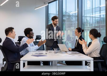 A diverse group of businesspeople clapping and smiling during a meeting in a modern office space with natural light. Stock Photo