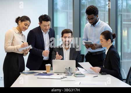 A diverse group of business professionals collaborates around a table in a modern office setting, focused on a laptop and documents. Stock Photo