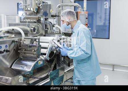Caucasian scientist in blue lab uniform configure manufacture machine with shafts and control panel, checking readings, making notes Stock Photo