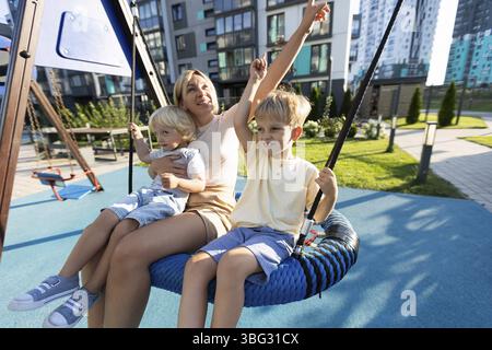 A mother shares joyful moments with her two young sons as they swing together on a playground. The bright day creates a cheerful atmosphere as laughte Stock Photo