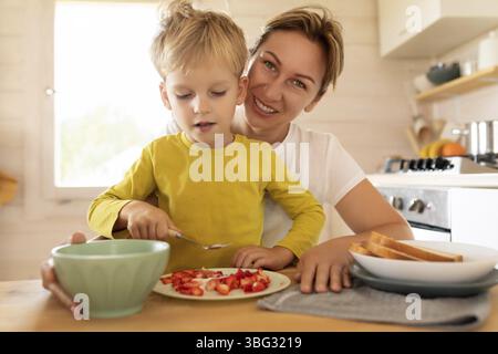 Cheerful Caucasian mother and little son are sitting in the kitchen and preparing breakfast Stock Photo