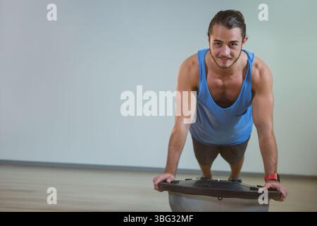 Man performing push-up on balance trainer wearing tank top shorts fitness tracker in workout studio Stock Photo