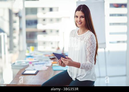 Office woman holding tablet while sitting on desk edge at office with sticky notes, copy space Stock Photo