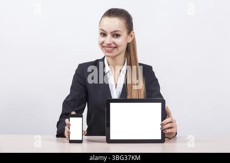 Stylish young business woman in a business suit sitting at a desk and is holding tablet and phone screen forward Stock Photo