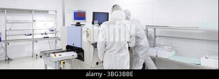 Two factory workers in white lab suits, working with some modern equipment, looking into the monitors in a clean room. panoramic picture Stock Photo