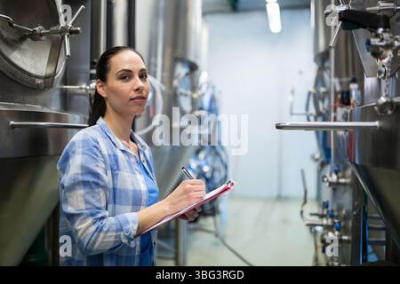 Female brewery technician standing between steel tanks in hall taking notes with clipboard and pen Stock Photo