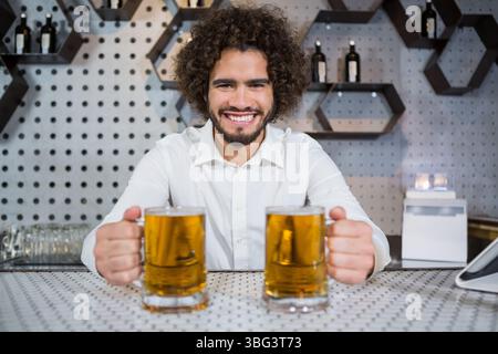 Man holding two frothy beer mugs and leaning behind perforated bar counter with honeycomb shelves Stock Photo