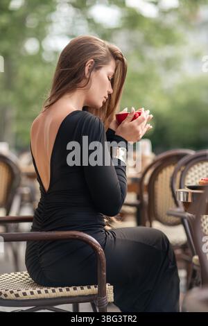 Woman Coffee Relaxation: Woman in black dress enjoys coffee at outdoor cafe, relaxing moment during the daytime. Stock Photo