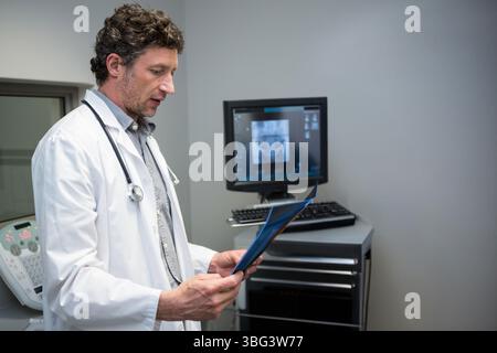 Middle-aged doctor holding blue x-ray folder with stethoscope and examining monitor in clinic Stock Photo