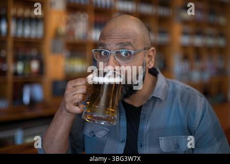Indian man wearing eyeglasses lifting clear glass mug with beer foam while sitting at pub counter Stock Photo