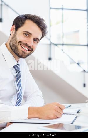 Male wearing shirt and tie writing in notebook beside tablet at office desk near window panes Stock Photo