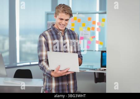 Man holding laptop smiling at screen in modern office with glass board and sticky notes Stock Photo