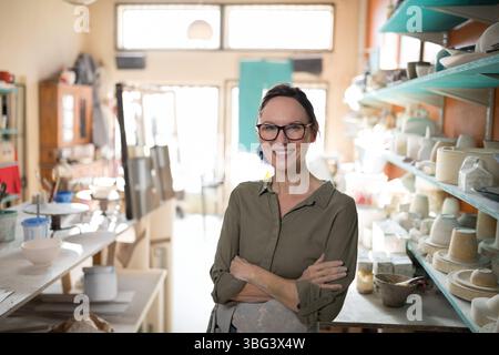 Woman wearing clay-stained apron standing in pottery workshop beside potter's wheel and clay bowls Stock Photo