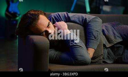 Man laying down hugging pillow watching TV, heartbroken by upsetting movie plot. Sad viewer lounged on couch in RGB illuminated apartment, upset by depressing television show scene, camera A Stock Photo
