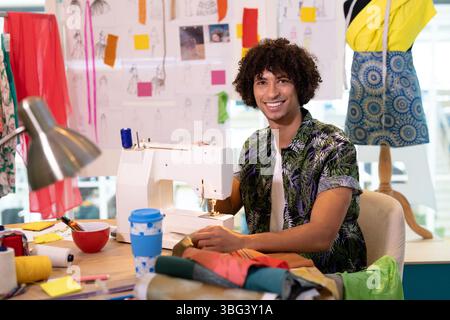 Adult male fashion designer sitting in workshop, using sewing machine with spools of thread Stock Photo