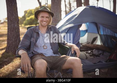 Male camper relaxing in collapsible camping chair at forest clearing with dome tent and backpack Stock Photo