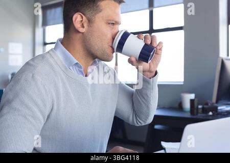 Man in knit sweater sipping takeaway coffee while working on laptop with papers at office desk Stock Photo