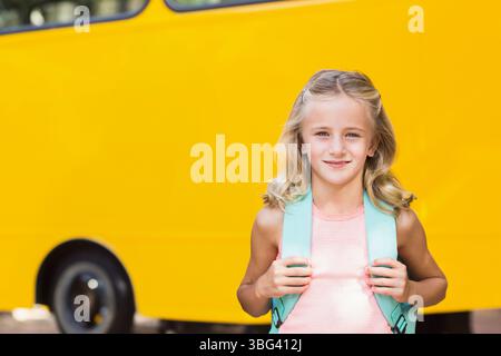 Smiling girl standing before yellow school bus grasping mint green backpack straps on sunny street Stock Photo