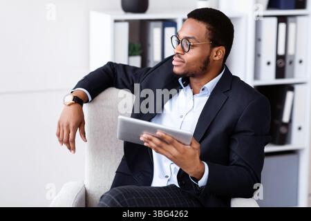 Thoughtful black businessman sitting in chair, using digital tablet Stock Photo