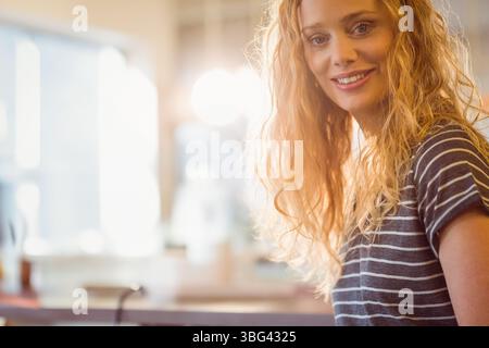 Adult woman smiling while leaning on kitchen countertop with utensils and window light, copy space Stock Photo