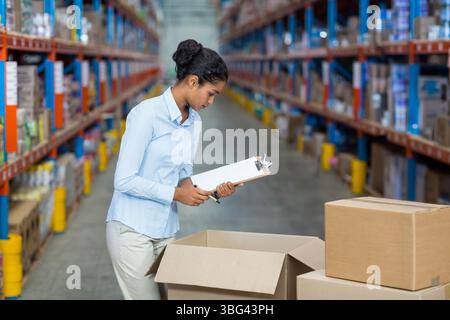 Woman holding clipboard and pen inspecting open cardboard box on pallet in warehouse aisle Stock Photo
