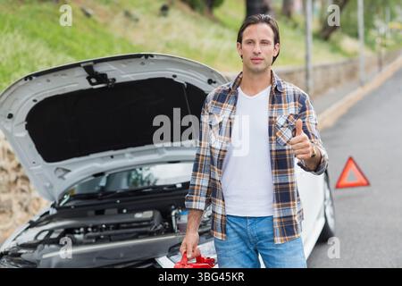 Middle-aged man standing beside sedan on road near red triangle, holding jumper cables, copy space Stock Photo