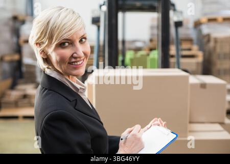 Middle-aged woman wearing blazer standing in warehouse holding clipboard near forklift, copy space Stock Photo