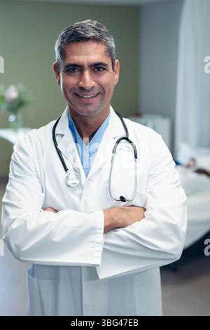 Senior male doctor standing in hospital room wearing lab coat and stethoscope beside patient bed Stock Photo