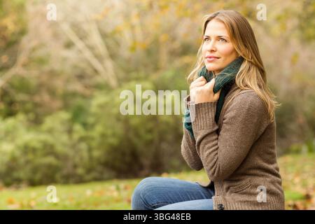 Woman sitting in park wearing cardigan and green scarf, gazing left amid fallen leaves, copy space Stock Photo