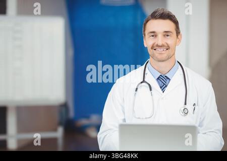 Male doctor sitting at clinic desk wearing stethoscope and using laptop near whiteboard Stock Photo