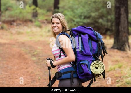 Female hiker standing on pine trail smiling carrying backpack with sleeping mat and trekking poles Stock Photo