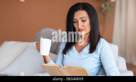 Mature lady reading book and holding cup of coffee Stock Photo