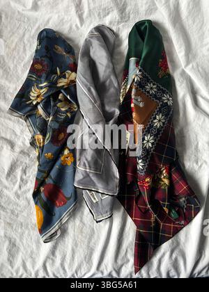 Three thrifted silk scarves—floral, striped, and holiday-patterned—are artfully draped on white linen, showcasing luxurious prints and textures. Stock Photo