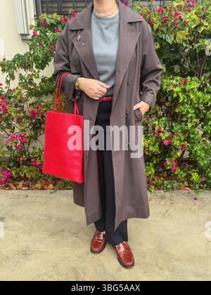 A woman in a brown trench coat, gray sweater, black trousers, red belt and red leather tote stands before pink bougainvillea. Stock Photo