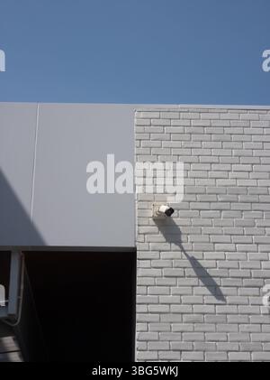 A modern building facade featuring a security camera mounted on a white brick wall. The clear blue sky adds depth to the architectural design. Stock Photo