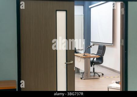 A modern office meeting room visible through a partially open door, featuring a sleek table, rolling chair, and a large screen. Stock Photo